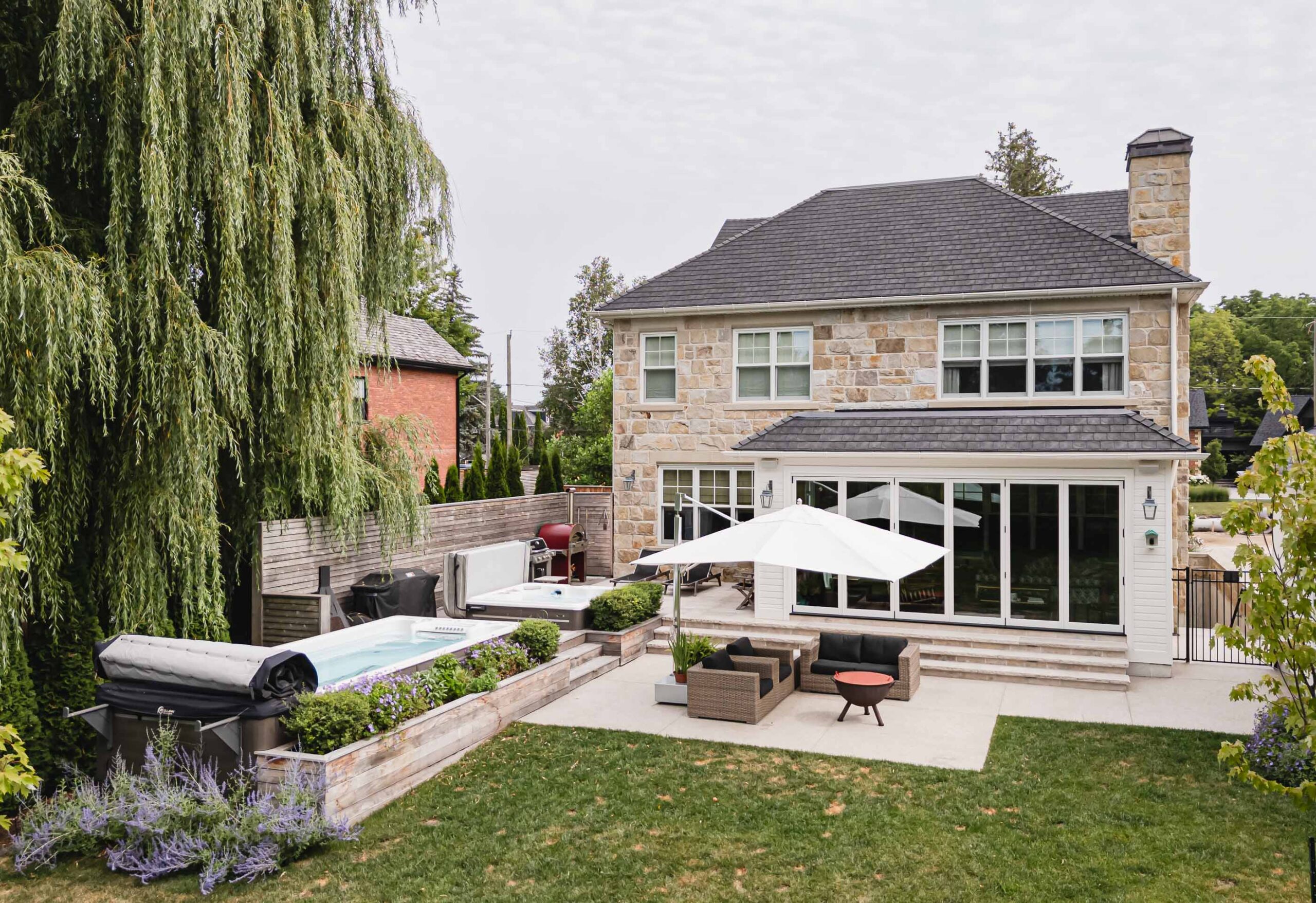 Backyard of an English-style stone house with swim spa, hot tub and lounge area, built by a Collingwood custom home builder