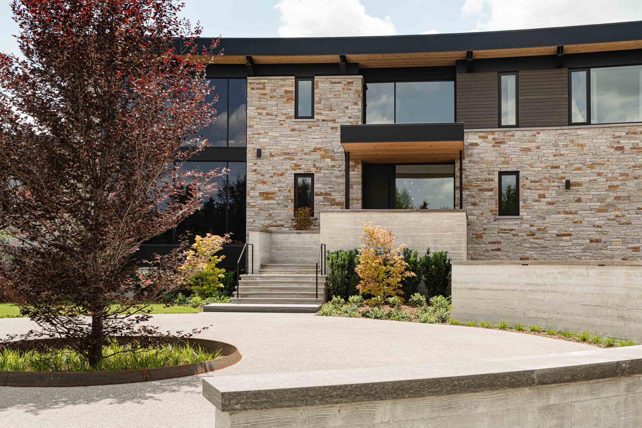 Front detail of a modern custom home with curved design, board formed concrete details, stone façade, and landscaped grounds– built by a Collingwood custom home builder