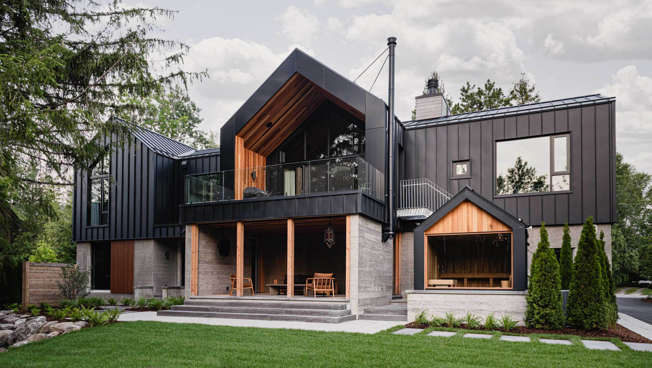Rear elevation of a nordic-inspired chalet with modern black siding, board formed concrete walls, screened sitting area and attached sauna
