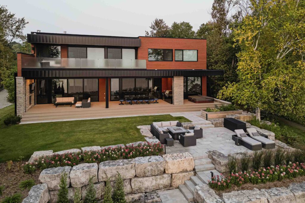 Exterior of a contemporary, waterfront home with extensive landscaping along the rocky shoreline and multiple outdoor seating areas down to the waters edge. Built by Blake Farrow Project