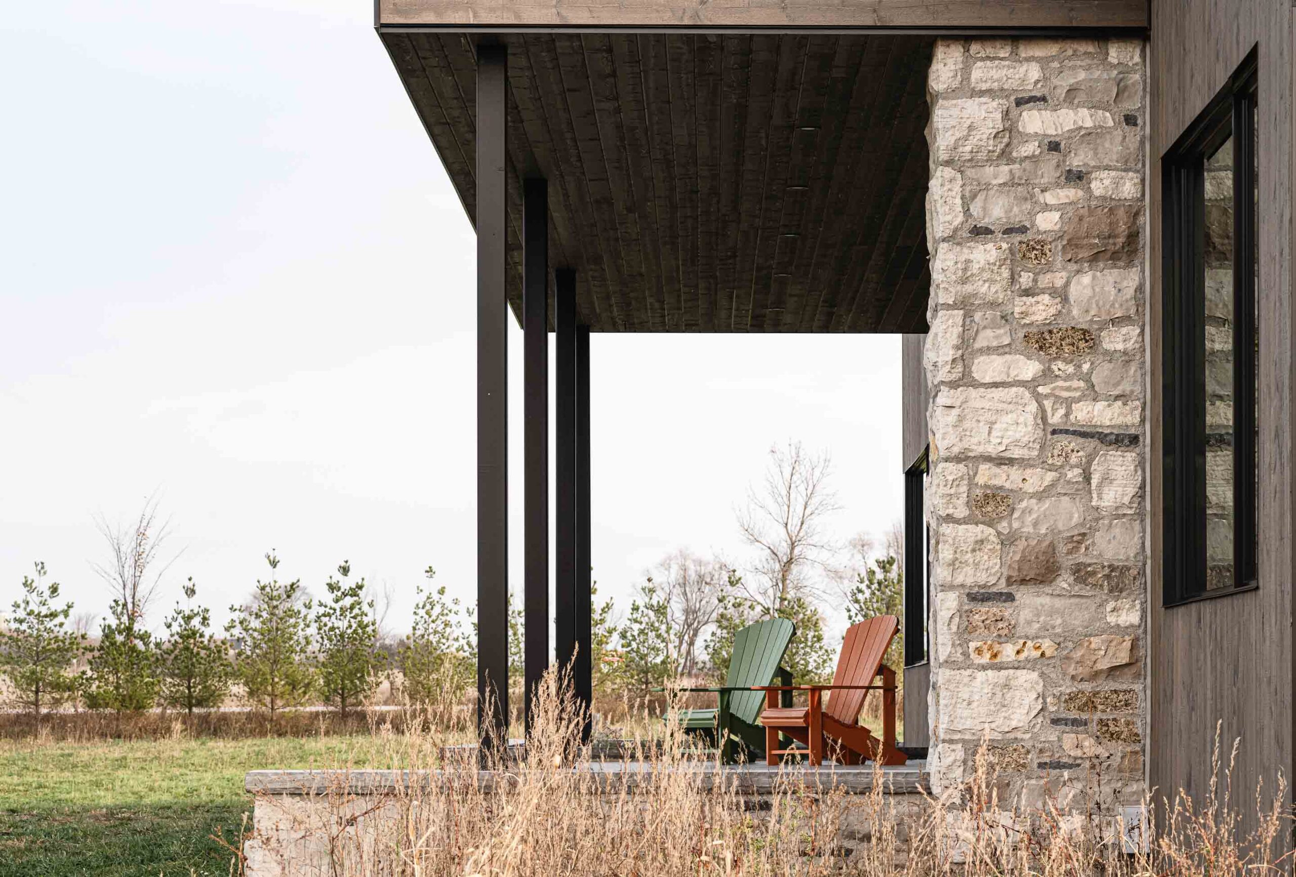 two Muskoka chairs sit on the front porch of a contemporary farmhouse with stone and metal details