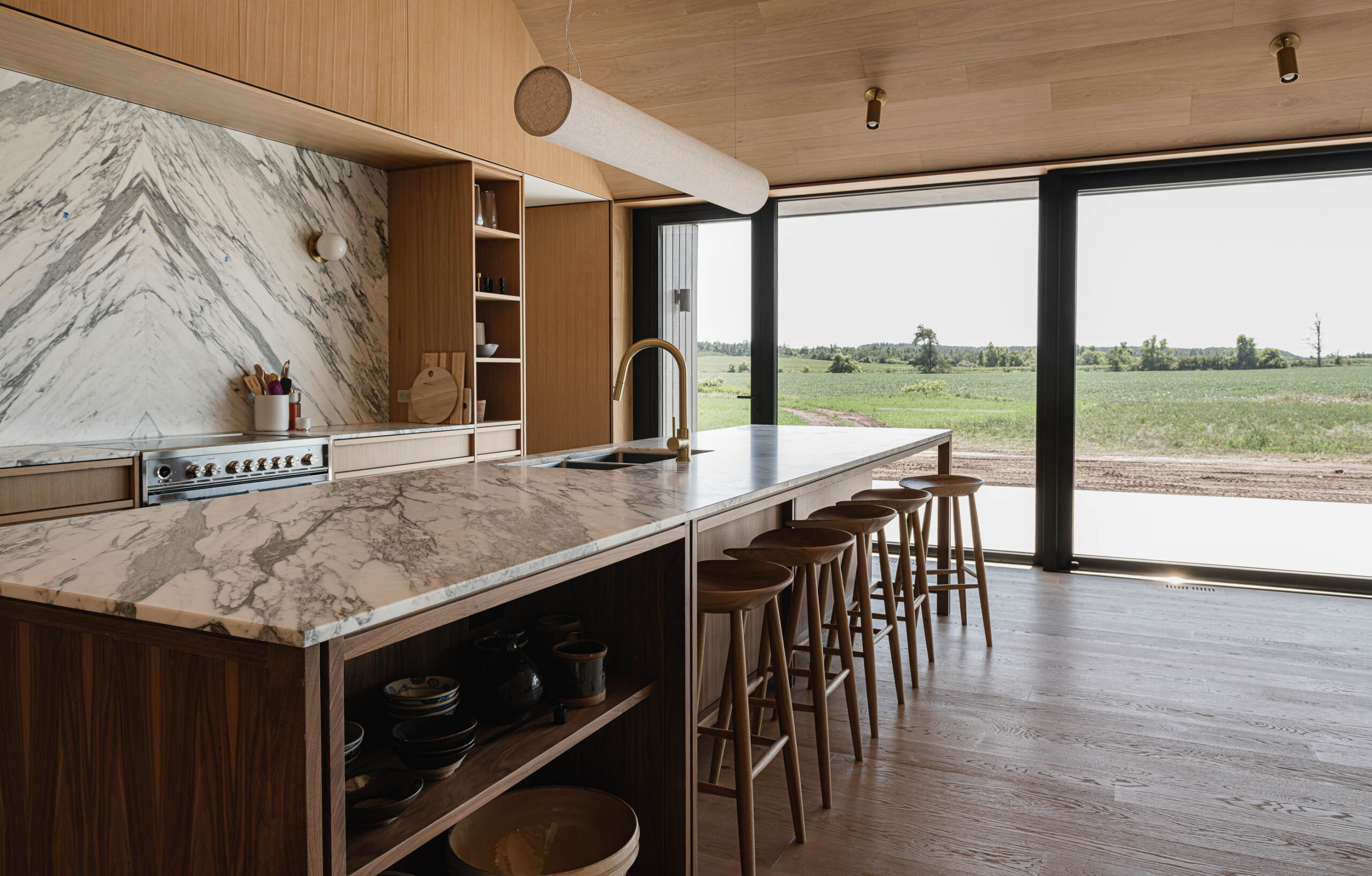 Kitchen island in a modern barn home with bookmatched quartz backsplash, custom walnut cabinetry and floor-to-ceiling glazing overlooking the surround country side