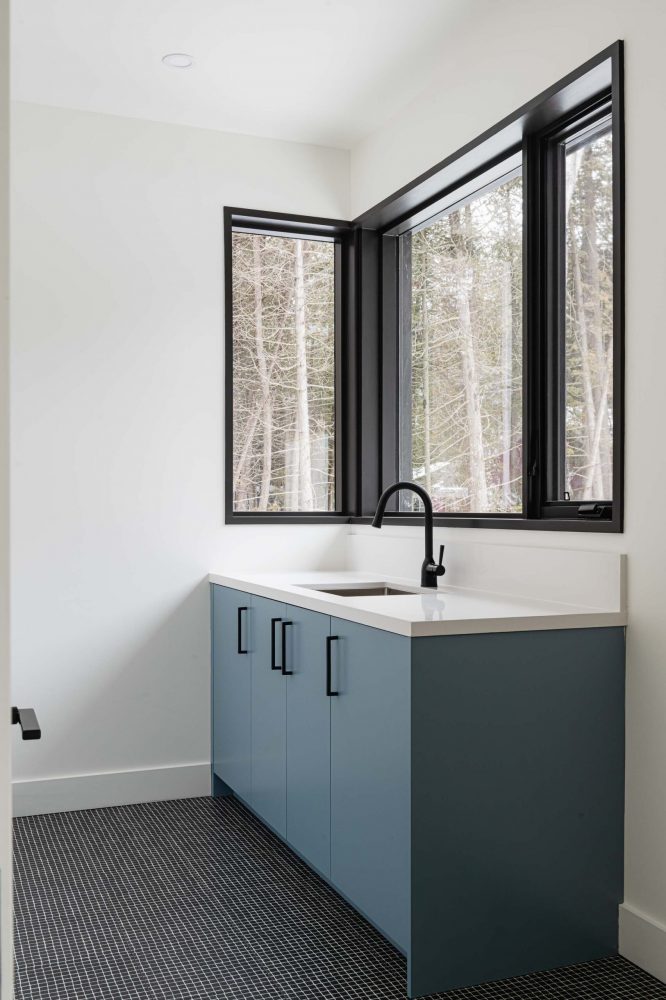 21-Modern-Waterfront a clean, modern laundry room with light blue cabinetry, black geometric fllor tiles and windows looking into the forest