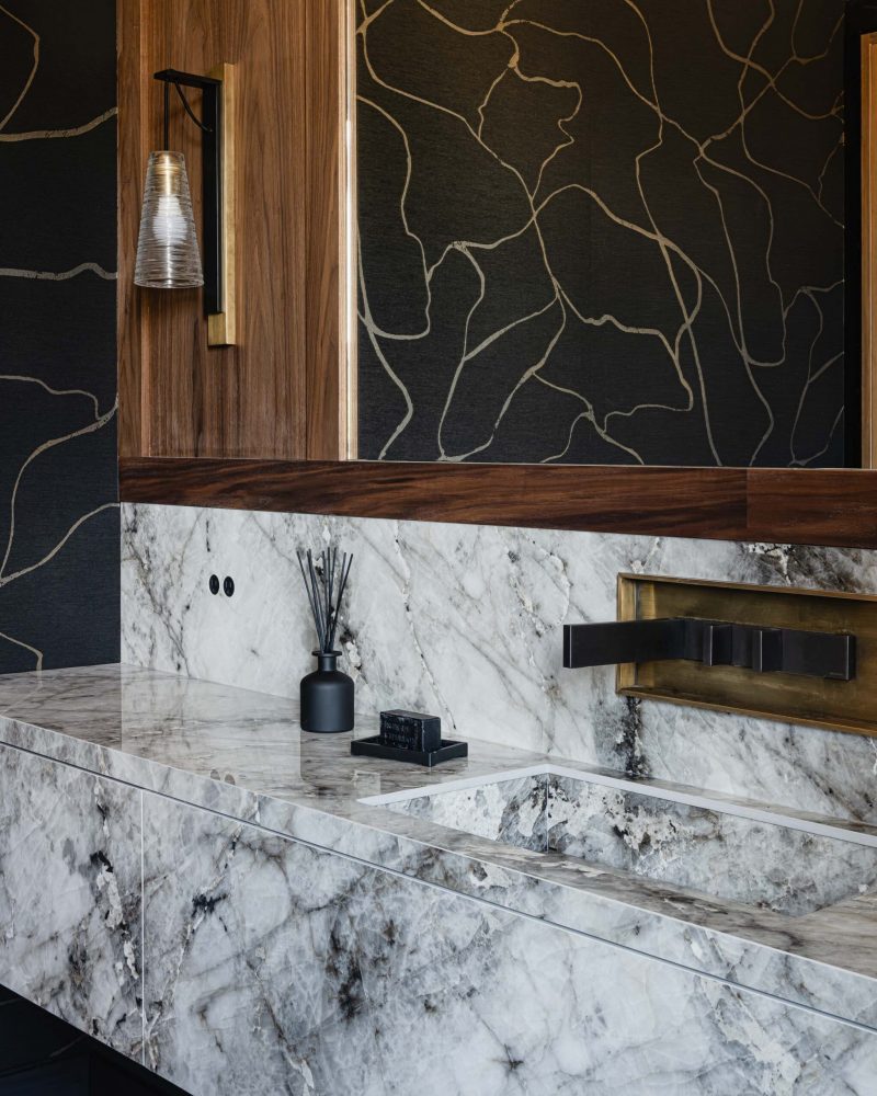 Contemporary Blue Mountain bathroom detail with sleek marble countertops and backsplash, modern brass faucet, and textured, black and gold wallpaper