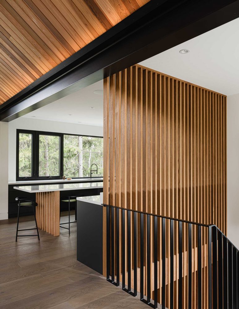 Modern alpine chalet kitchen with slat wood divider wall, expansive forest views, and exposed steel beam with recessed up lighting